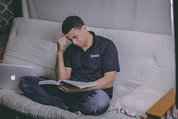 A student relaxing on a sofa, studying with a book and using a laptop indoors. HD wallpaper