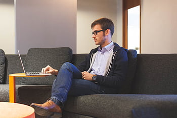 A young man in casual attire working remotely on a laptop on a couch indoors. HD wallpaper