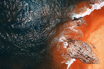 A stunning aerial shot of a vibrant seashore with waves crashing against rocky formations and orange sands. HD wallpaper