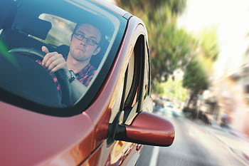 A man driving fast through a city street in a red car, showcasing motion and focus. HD wallpaper