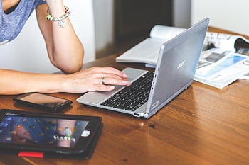 Woman working remotely on a laptop with mobile devices on a wooden desk. HD wallpaper