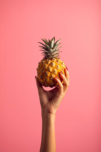 A hand with polished nails holds a ripe pineapple against a vivid pink backdrop, embodying tropical freshness. HD phone wallpaper
