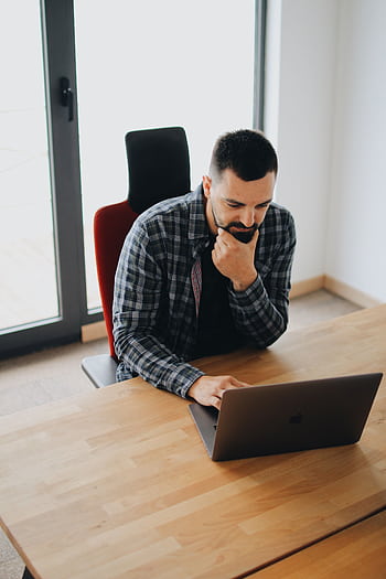 Young man with beard working on laptop at desk in a modern office setting. HD phone wallpaper