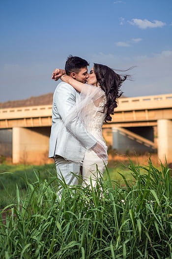 A young couple embraces and kisses in a field near a bridge, showcasing love and romance. HD phone wallpaper