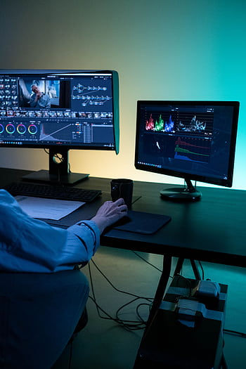 A video editor works on dual monitors in a modern setup, focusing on color grading and editing visuals. HD phone wallpaper