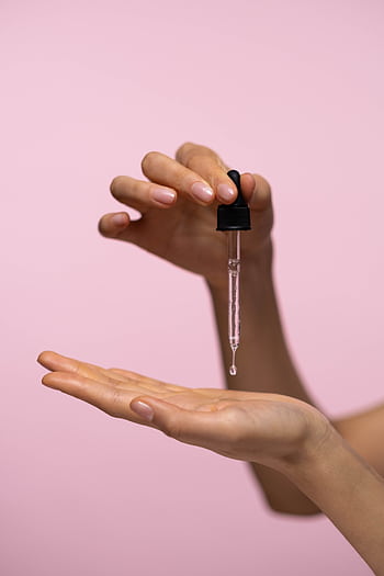 Close-up of hands with dropper applying face serum against a pink background for skincare routine. HD phone wallpaper