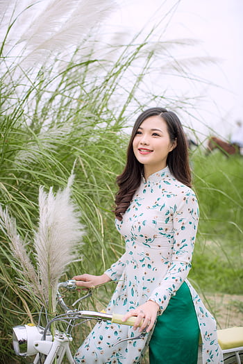 A young woman in a floral dress smiles beside a bicycle in a grassy field, embracing nature. HD phone wallpaper