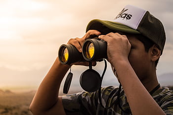 A young man wearing a cap uses binoculars to observe the scenic landscape outdoors during daylight. HD wallpaper