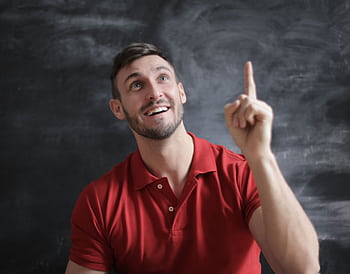 Happy young man in a red polo shirt smiling and pointing upwards in front of a blackboard. HD wallpaper