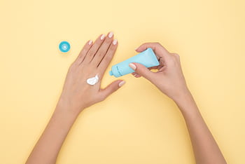 Close-up of a woman's hands applying cream on a pastel yellow background. Ideal for beauty and skincare themes. HD wallpaper
