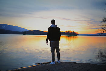 A man stands by a tranquil lake at sunset, reflecting the serene sky. HD wallpaper