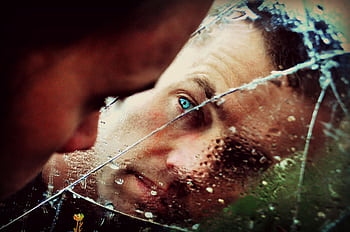 A man with green eyes peers into a cracked, wet mirror, conveying solitude and thought. HD wallpaper