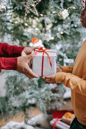 Hands exchanging a wrapped gift in front of a decorated Christmas tree. HD phone wallpaper