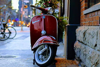 A classic red Vespa scooter parked beside a brick wall in an urban street setting. HD wallpaper