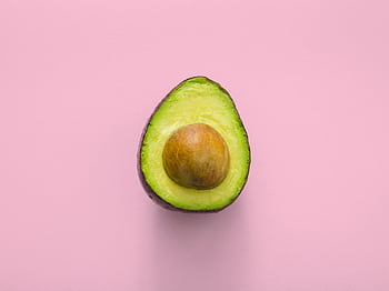 Fresh avocado fruit cut in half, showcasing seed, on a pastel pink background. HD wallpaper