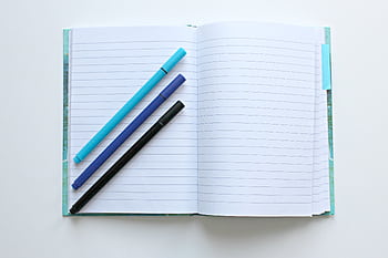 Open lined notebook with colorful pens on a white surface, perfect for note-taking and office tasks. HD wallpaper