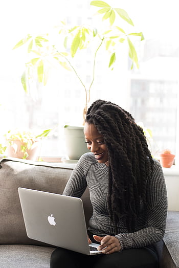 Black woman sitting on a sofa, smiling while working on a laptop indoors. HD phone wallpaper