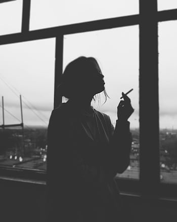 A moody black and white silhouette of a woman smoking, captured by a window with an urban backdrop. HD phone wallpaper