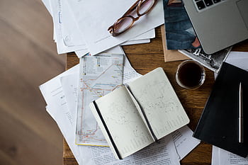 Flat lay of a workspace with coffee, maps, laptop, and documents on a wooden desk. HD wallpaper