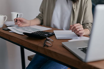 A woman manages finances at home, using a laptop and calculator on a wooden desk. HD wallpaper