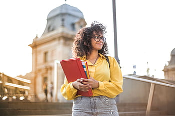 Smiling female student with curly hair and glasses holding books on campus steps. HD wallpaper