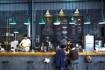 A bustling café with a modern industrial design, featuring a barista making coffee and patrons enjoying drinks. HD wallpaper