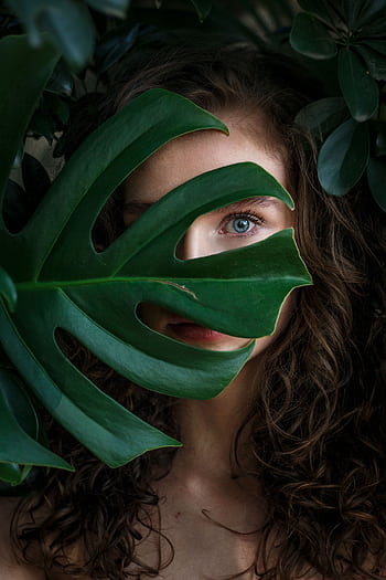 A beautiful woman peeks through lush green leaves in this captivating portrait. HD phone wallpaper