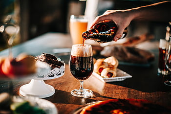 Sunlit scene with hand pouring dark beer into a glass. Includes baked goods and desserts on table. HD wallpaper