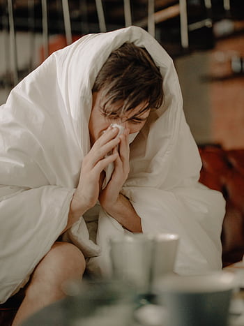 A young man wrapped in a blanket sneezes, indicating illness or cold symptoms. HD phone wallpaper