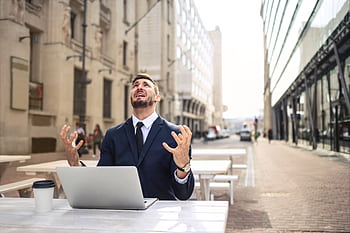 Businessman in suit showing frustration at outdoor table with laptop and coffee. HD wallpaper