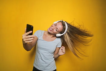 Cheerful woman wearing heads, enjoying music and taking a selfie against a vibrant yellow background. HD wallpaper