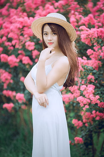 A beautiful woman in a white dress and straw hat poses gracefully in a vibrant garden full of pink flowers. HD phone wallpaper
