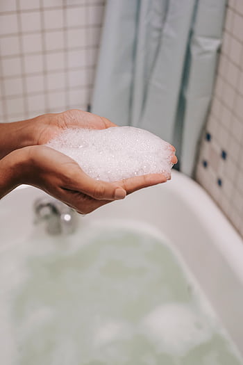 Close-up of hands holding bubble foam above a filled bathtub, creating a relaxing bath ambiance. HD phone wallpaper