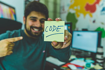 A smiling programmer pointing to a sticky note with 'code' written on it in an office setting. HD wallpaper