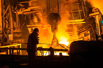A worker operates machinery in a factory with molten metal and intense flames. HD wallpaper