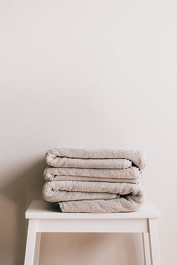 Stacked beige towels on a white stool in a minimalist bathroom setting, conveying freshness and comfort. HD phone wallpaper