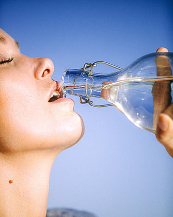 A woman drinking water from a glass bottle against a clear blue sky, emphasizing hydration. HD phone wallpaper