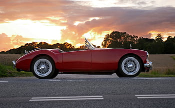 Vintage red convertible car parked on a country road at sunset with dramatic skies. HD wallpaper