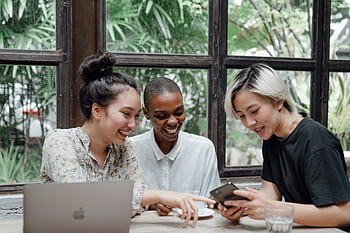Three friends enjoy laughter and connection over coffee, showcasing diverse togetherness indoors. HD wallpaper