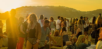A lively gathering of people enjoying a sunset party on a beach in Mykonos, Greece. HD wallpaper