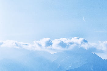 A tranquil scene of mountains enveloped by clouds with a crescent moon in a bright blue sky. HD wallpaper