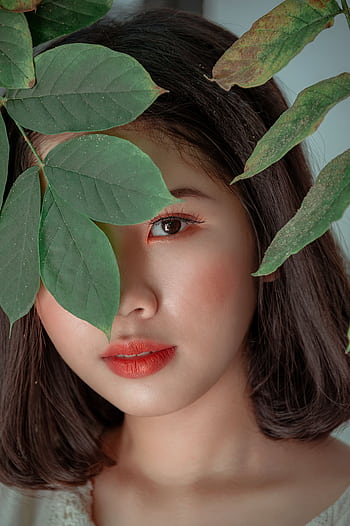 Close-up portrait of a woman peeking through green leaves, showcasing beauty and nature. HD phone wallpaper