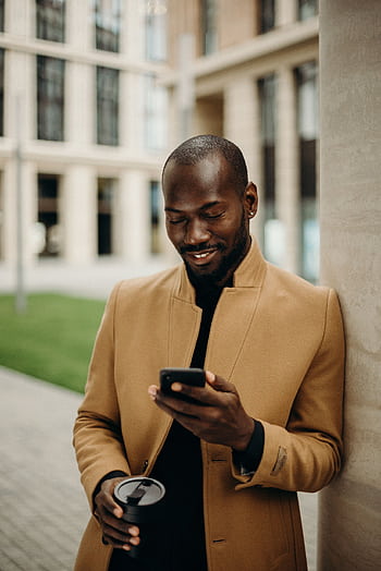 Relaxed man in coat smiles while using smart and holding coffee outside. HD phone wallpaper