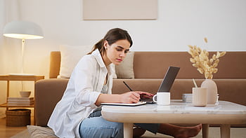 A young woman focuses on her studies while using a laptop in a cozy, modern living room. HD wallpaper