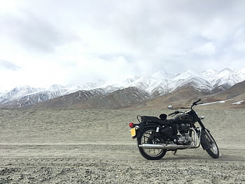 A vintage motorcycle parked on a gravel road with snowcapped mountains in the background, under a cloudy sky. HD wallpaper