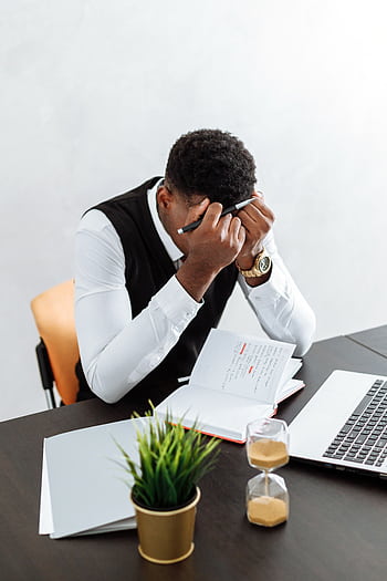 A businessman overwhelmed working indoors with a notebook, laptop, and hourglass. HD phone wallpaper