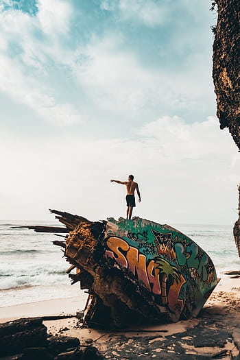 A man stands atop a graffiti-painted shipwreck on a beach in Bali, Indonesia, capturing moments of adventure and exploration. HD phone wallpaper