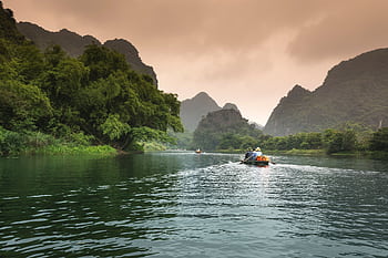 Two people rowing a boat through a serene river surrounded by lush greenery and mountains. HD wallpaper