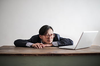 Asian businessman in glasses, resting head on desk, looking at laptop in office setting. HD wallpaper