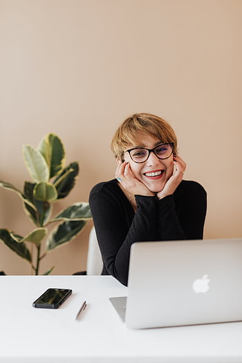 Cheerful woman in eyeglasses and black sweater smiling while working at a desk with a laptop indoors. HD phone wallpaper